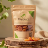 High-quality organic dried apricots in eco-friendly brown kraft packaging with a transparent window revealing plump golden-orange fruit, displayed on rustic stacked wooden slices surrounded by fresh green leaves and soft natural sunlight, branded Organica The Organic Store.