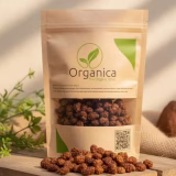 A premium resealable beige pouch of Organica Organic Dried Mulberries displayed on a rustic wooden platform. The packaging features a green leaf logo and clear window showing brown sun-dried mulberries inside. Loose dried mulberries are scattered artistically on the textured wood surface in the foreground, set against a warm, blurred natural background with soft daylight lighting.