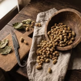 A premium resealable beige pouch of Organica Organic Dried Mulberries displayed on a rustic wooden platform. The packaging features a green leaf logo and clear window showing brown sun-dried mulberries inside. Loose dried mulberries are scattered artistically on the textured wood surface in the foreground, set against a warm, blurred natural background with soft daylight lighting.