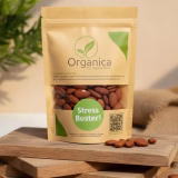 Organica Premium Roasted Almonds brown paper pouch standing centrally on stacked rustic wooden boards. The packaging features a green leaf logo and prominent "Stress Buster!" text. Fresh, crunchy baked almonds are scattered on the wood surface, illuminated by warm, natural sunlight with a blurred organic green background
