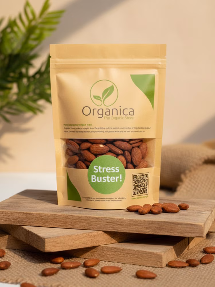 roasted-almonds-premium-natural-organica Organica Premium Roasted Almonds brown paper pouch standing centrally on stacked rustic wooden boards. The packaging features a green leaf logo and prominent "Stress Buster!" text. Fresh, crunchy baked almonds are scattered on the wood surface, illuminated by warm, natural sunlight with a blurred organic green background