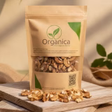 Organica The Organic Store premium walnut kernels (Akhrot Giri) packaging standing on a rustic wooden platform, surrounded by scattered shelled walnuts and soft green leaves in warm sunligh